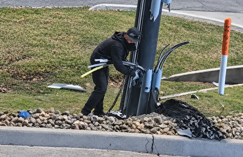 A worker in dark clothing repairs a damaged roadside utility pole amid scattered debris and exposed cables, creating a tense scene of infrastructure damage.